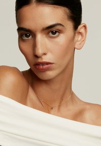 Collar de oro con un colgante de luna creciente, usado sobre un hombro descubierto. La modelo tiene el cabello oscuro, maquillaje neutro y una blusa blanca sin hombros.