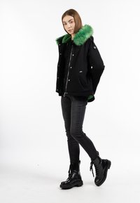 Giubbotto nero con cappuccio foderato in pelliccia verde, chiusura con zip e bottoncini a pressione. Modello che indossa jeans scuri e stivali neri con lacci.