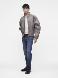 Jeune homme portant une veste en cuir grise, une chemise blanche, un jean bleu et des chaussures noires, debout devant un fond blanc uni.