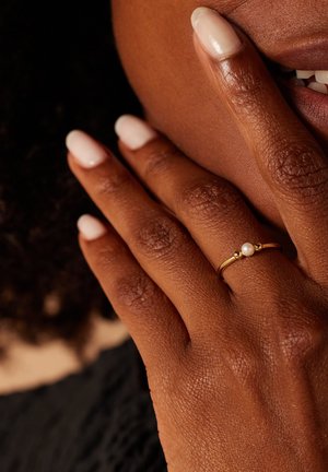 Gouden ring met een gecentreerde parel en twee kleine accentbedels. Hand met mediumbruine huid en korte, neutraal gekleurde nagels die op het gezicht rusten.
