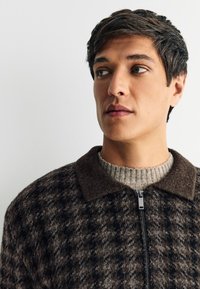 Veste zippée marron à carreaux avec une texture douce, dotée d'un col sombre et d'un pull léger côtelé en dessous. Le visage du modèle est partiellement visible.