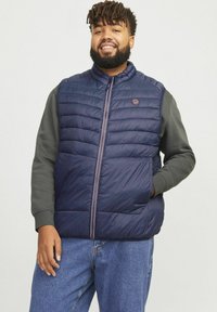 Marineblauer Puffy-Weste mit horizontalen Nähten, frontalem Reißverschluss und kleinem Logo. Kombiniert mit einem grauen Langarmshirt und Jeans.