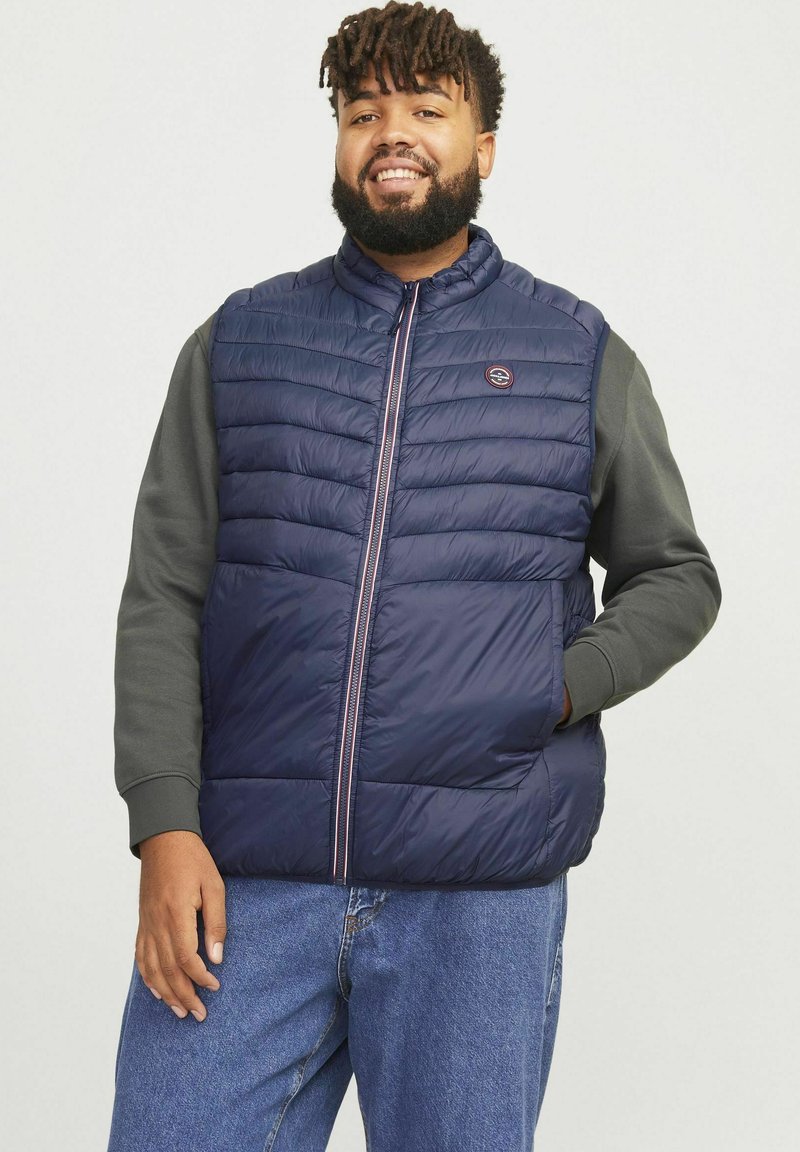 Marineblauer Puffy-Weste mit horizontalen Nähten, frontalem Reißverschluss und kleinem Logo. Kombiniert mit einem grauen Langarmshirt und Jeans.