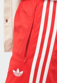 Rote sportliche Shorts mit weißen Streifen, Reißverschlusstaschen und einem weißen, aufgestickten Logo unten links. Hergestellt aus einem glatten Stoff.