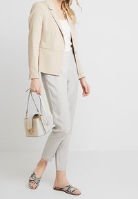 Beige blazer, vit topp, ljusgrå byxor och mönstrade slip-on skor. Bärande en liten beige handväska. Släta texturer, skräddarsydd passform.