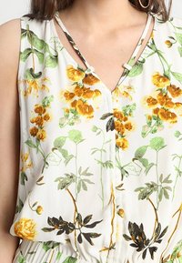 Blouse sans manches imprimée florale avec un fond blanc, des fleurs orange et vertes ainsi que des motifs de feuilles. Design à encolure en V avec des bretelles décoratives.