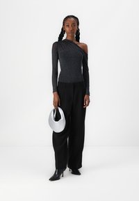 Schwarzes Top mit einem Schulterträger und strukturiertem, schimmerndem Finish, kombiniert mit lockeren schwarzen Hosen und eleganten Stiefeletten. Hält eine silberne Handtasche.