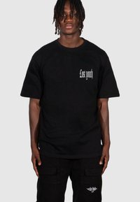 Sort bomulds T-shirt med teksten "Lost youth" i hvid gotisk skrift på venstre bryst, parret med sorte cargo-bukser.