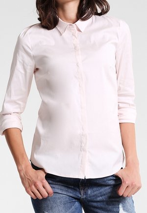 Kobieta ubrana w jasnoróżową koszulę na guziki z podwiniętymi rękawami i niebieskie przetarte dżinsy, stoi z rękami w kieszeniach.