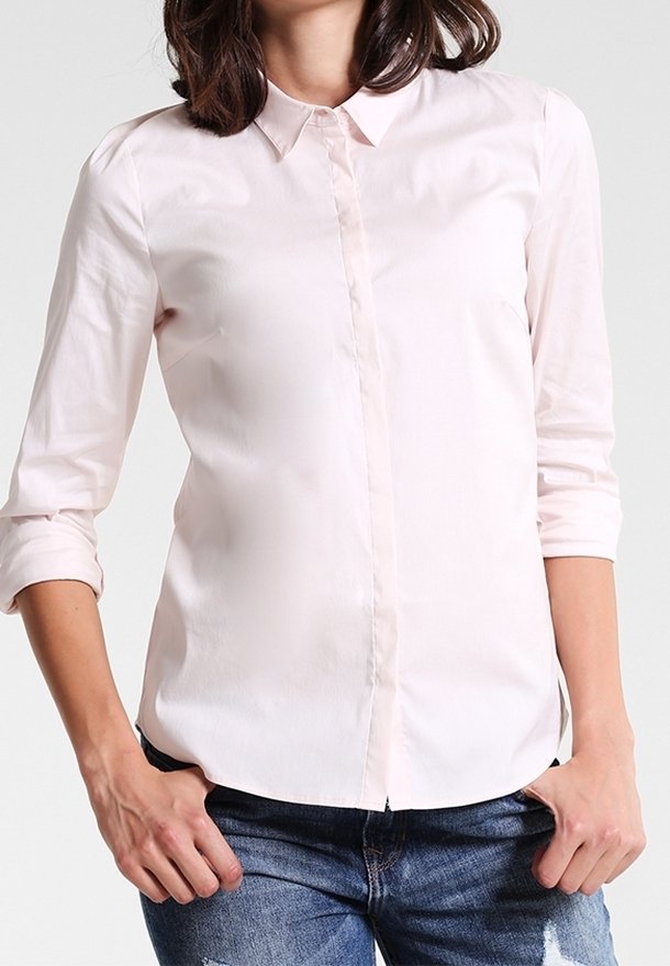 Femme portant une chemise rose pâle à boutons avec les manches retroussées et un jean bleu déchiré, debout les mains dans les poches.