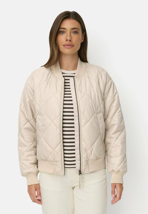 Frau trägt eine hellbeige gesteppte Bomberjacke über einem braun-weiß gestreiften Hemd und weißen Hosen, steht vor einem einfarbigen Hintergrund.
