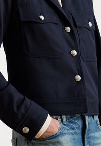 Primo piano di una persona che indossa una giacca blu navy con bottoni argentati e tasche a patta, abbinata a jeans blu in denim e una camicia beige.