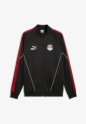 Veste de survêtement Puma noire avec des bandes rouges à motifs sur les manches, passepoil blanc, texte "KING" et emblème avec un ballon de football et un drapeau sur la poitrine.
