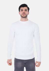 Maglione bianco a treccia lavorato a maglia con colletto e polsini a costine; design aderente; texture morbida; indossato sopra jeans scuri.