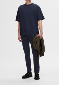 Laivastonsininen ylipitkä t-paita lyhyillä hihoilla, yhdistettynä tummiin housuihin. Pitelee oliivinvihreää teksturoitua neuletta yhdessä kädessä. Mustat kengät yksityiskohdilla.