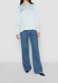 Camicia azzurra a maniche lunghe con colletto abbottonato, abbinata a jeans blu in denim a gamba larga e scarpe nere con punta appuntita.