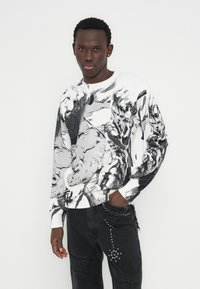 Μπλούζα πλεκτή ασπρόμαυρη με καλλιτεχνικό σχέδιο, με ριμπ λαιμόκοψη και μακριά μανίκια, συνδυασμένη με μαύρο τζιν με σκισίματα και διακοσμητικά καρφιά.