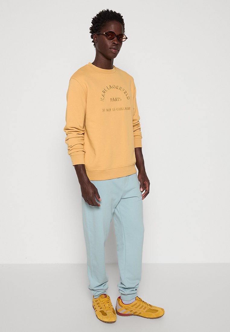 Gelbes Sweatshirt mit Logo, hellblaue Jogginghose und gelbe Sneakers. Das Outfit hat eine lockere Passform und ein einfaches Design. Braune Sonnenbrille getragen.
