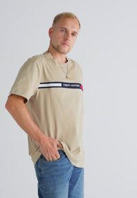 Beež lühikeste varrukatega t-särk, mille rinnal on horisontaalne tumesinine ja punane triip, millel on kaubamärk "TOMMY HILFIGER". Kandmisel sinised teksapüksid.