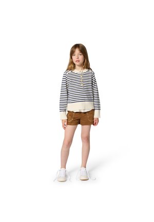 Mladé dievča oblečené v pruhovanom svetríku, hnedých šortkách s kovovými okami a bielych teniskách, stojace pred bielym pozadím.