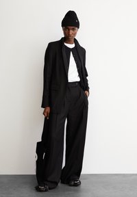 Schwarze taillierte Anzugjacke und hoch geschnittene, weit geschnittene Hose, über einem weißen Oberteil getragen, kombiniert mit einer schwarzen Mütze und fransigen Loafers.