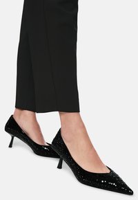 Schwarze spitze Pumps mit einem strukturierten Obermaterial, das mit kleinen Pailletten bedeckt ist, und einem niedrigen Kegelabsatz. Kombiniert mit eng anliegenden schwarzen Hosen.