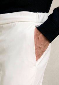 Fehér szövetnadrág sima felülettel, oldalsó zsebekkel és rugalmas derékkal. Egy sötét pulóver félig látható fölötte.