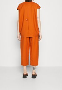 Oranžový dvoudílný outfit, který zahrnuje top s krátkým rukávem a detaily knoflíku na zádech a kalhoty širokého střihu. Hladká textura, zkrácená délka.