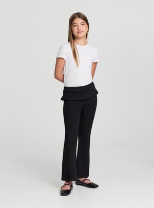 Ragazza giovane in piedi con le mani dietro la schiena, indossa una maglietta bianca a maniche corte, pantaloni neri con dettaglio a strati in vita e scarpe nere con cinturino.