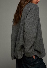 Frau mit langen braunen Haaren, die einen lockeren grauen Strickpullover und dunkle Jeans trägt, seitlich steht und eine Hand in der Tasche hat, vor einem schlichten Hintergrund.