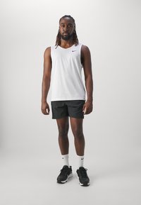 T-shirt de sport sans manches blanc avec logo noir, associé à un short noir. Modèle portant des baskets noires et des chaussettes blanches. Fond neutre.