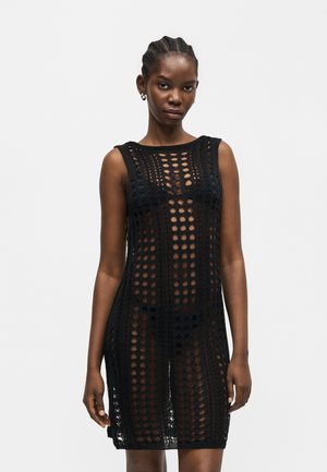 Femme portant une robe noire en crochet sans manches avec des motifs circulaires, debout devant un fond clair uni.