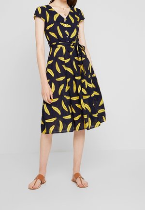 Kobieta ubrana w granatową sukienkę do kolan z żółtym nadrukiem bananów, krótkie rękawy, guziki, przewiązana w talii, brązowe płaskie sandały.