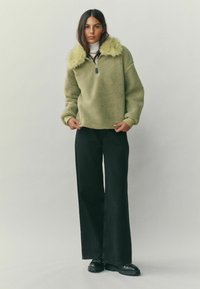 Heleroheline fliispusa kunstkarvaga kraega, esiosas poollukuga ja langevate õlgadega, kombineeritud laiade mustade pükstega.