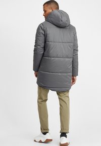 Grå polstret frakk med hette, med horisontalt sydd sektioner. Kombinert med lyse khaki bukser og hvite sneakers. Glatt tekstur.