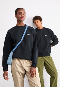 Must must, mille väikese logoga, koos beežide pükstega. Mudel hoiab helesinist kotti. Taustal on teine kuju mustas ujumistrikoos.