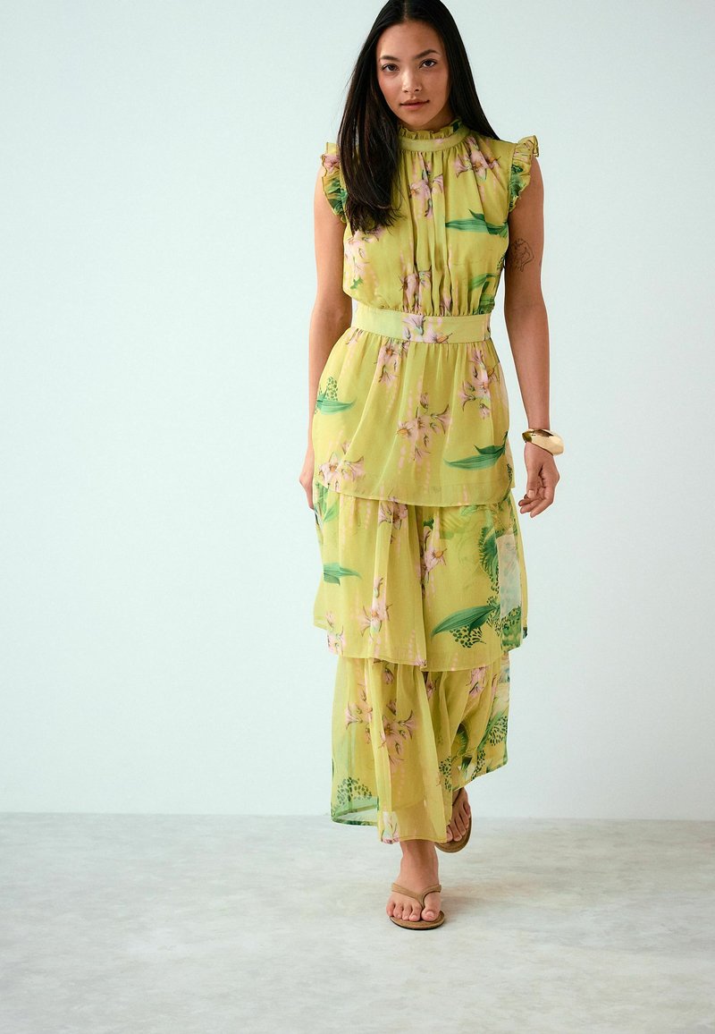 Femme aux longs cheveux noirs portant une robe maxi jaune à fleurs superposées et des sandales beiges, debout sur un sol gris clair contre un mur blanc.