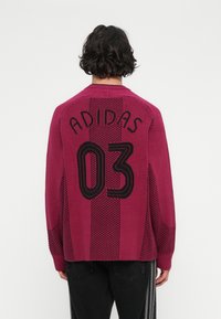 Vīrietis ar bordo Adidas džemperi, uz kura ir melns "ADIDAS 03" teksts, kombinēts ar melniem biksēm ar baltām vertikālām svītrām, parādīts no aizmugures.