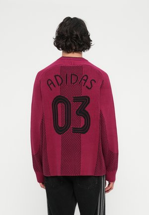 Moški nosi burgundski Adidas pulover z črnim napisom "ADIDAS 03", v kombinaciji s črnimi hlačami z belimi vertikalnimi črtami, prikazan od zadaj.