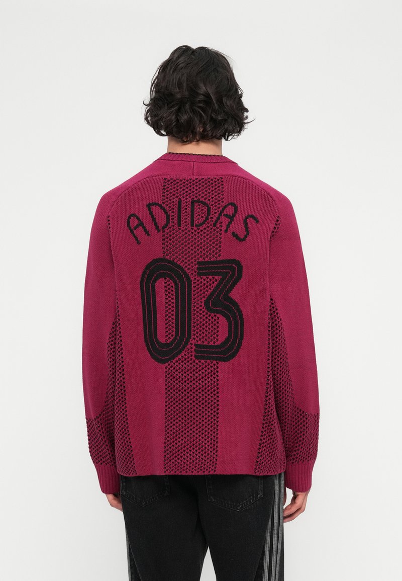 Vīrietis ar bordo Adidas džemperi, uz kura ir melns "ADIDAS 03" teksts, kombinēts ar melniem biksēm ar baltām vertikālām svītrām, parādīts no aizmugures.