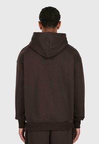 Persona vista di spalle che indossa una felpa con cappuccio marrone scuro e pantaloni coordinati contro uno sfondo chiaro e semplice.