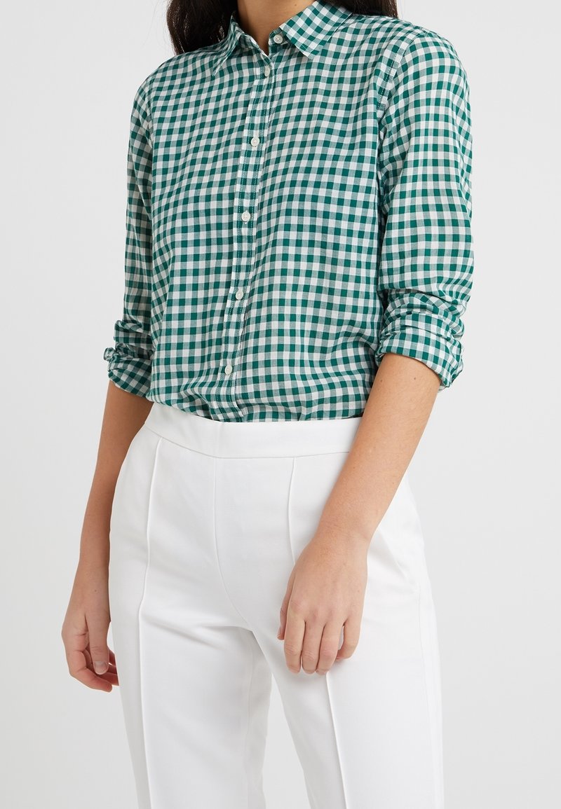 Femme portant une chemise vichy verte et blanche à boutons avec manches retroussées et pantalon blanc taille haute sur fond uni.