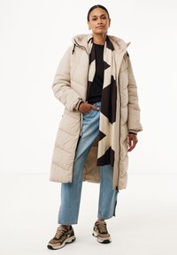 Lange beige pufferjas met een capuchon en rits aan de voorkant, gecombineerd met lichtblauwe jeans en bruine sneakers met een patroon. Aangevuld met een geometrische sjaal.