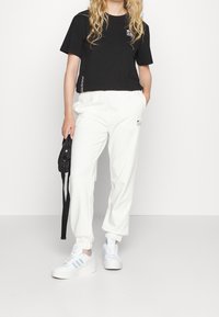 Frau in schwarzem Lacoste-T-Shirt, weißen Jogginghosen, weißen Adidas-Sneakers, die eine kleine schwarze Umhängetasche hält und vor einem weißen Hintergrund steht.