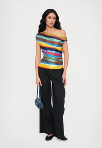 Multicolore off-shoulder top met verticale strepen in blauw, groen, bruin en geel. Gecombineerd met wijde zwarte broek en een kleine blauwe tas.