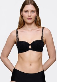 Reggiseno in pizzo nero con stecche, fantasie floreali, bordi scallop e dettaglio a chiave sul davanti. Spalline nere semplici con un design testurizzato.