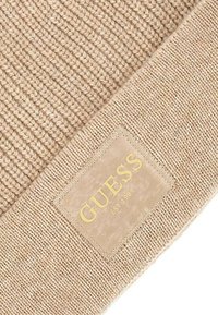 Tessuto lavorato a maglia beige con un motivo testurizzato. Presenta una toppa rettangolare in pelle marrone chiaro impresso con "GUESS" e "EST 1981."