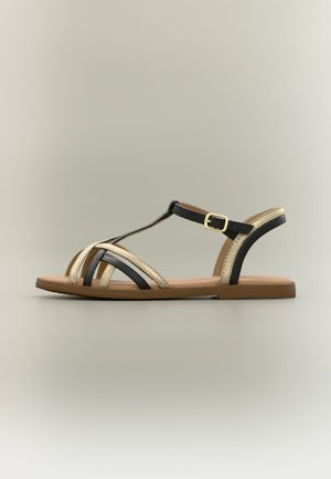 Sandalo basso con cinturini incrociati neri e beige, cinturino alla caviglia con fibbia dorata e suola marrone chiaro su sfondo neutro.
