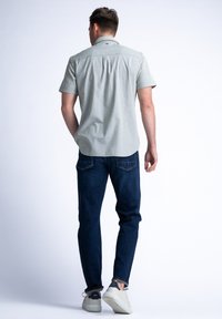 Camisa de botones de manga corta, verde claro con un patrón sutil, combinada con jeans azul oscuro y zapatillas blancas, vista desde atrás.