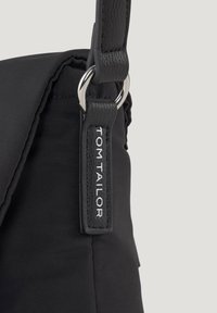 Primer plano de una bolsa de tela negra con correa de cuero y anillo plateado, que presenta una etiqueta negra cosida con la inscripción "TOM TAILOR" en letras blancas.